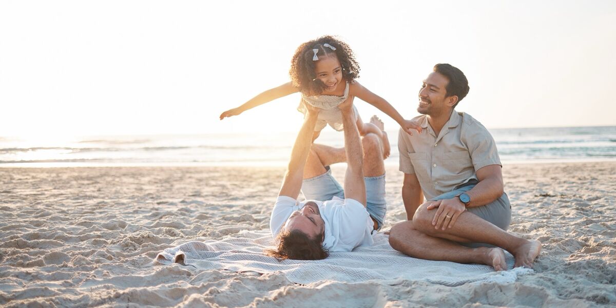 Zwei Väter mit ihrer Tochter am Strand, ein Vater hebt das Mädchen in die Luft. 