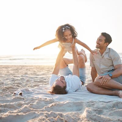 Zwei Väter mit ihrer Tochter am Strand, ein Vater hebt das Mädchen in die Luft.