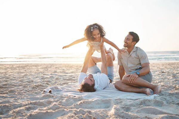 Zwei Väter mit ihrer Tochter am Strand, ein Vater hebt das Mädchen in die Luft.