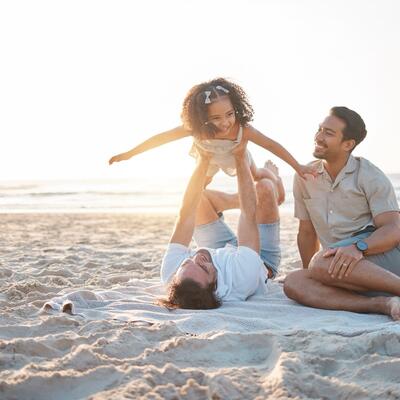 Zwei Väter mit ihrer Tochter am Strand, ein Vater hebt das Mädchen in die Luft.