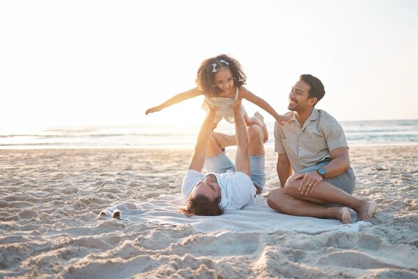 Zwei Väter mit ihrer Tochter am Strand, ein Vater hebt das Mädchen in die Luft.