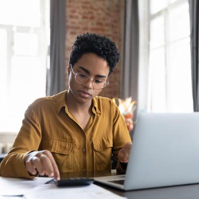 Junge Frau mit Brille sitzt am Laptop und rechnet etwas durch.