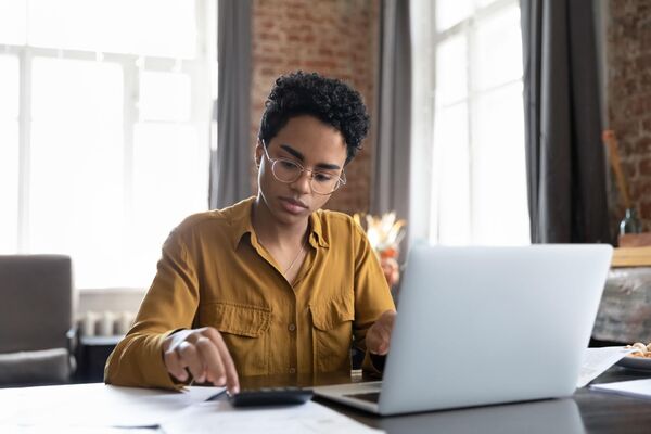 Junge Frau mit Brille sitzt am Laptop und rechnet etwas durch.