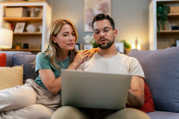Paar sitzt mit einem Computer auf dem Sofa und rechnet etwas nach.