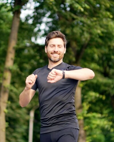 Attraktiver Mann über 40 joggt durch einen Wald und blickt auf seine Smartwatch.