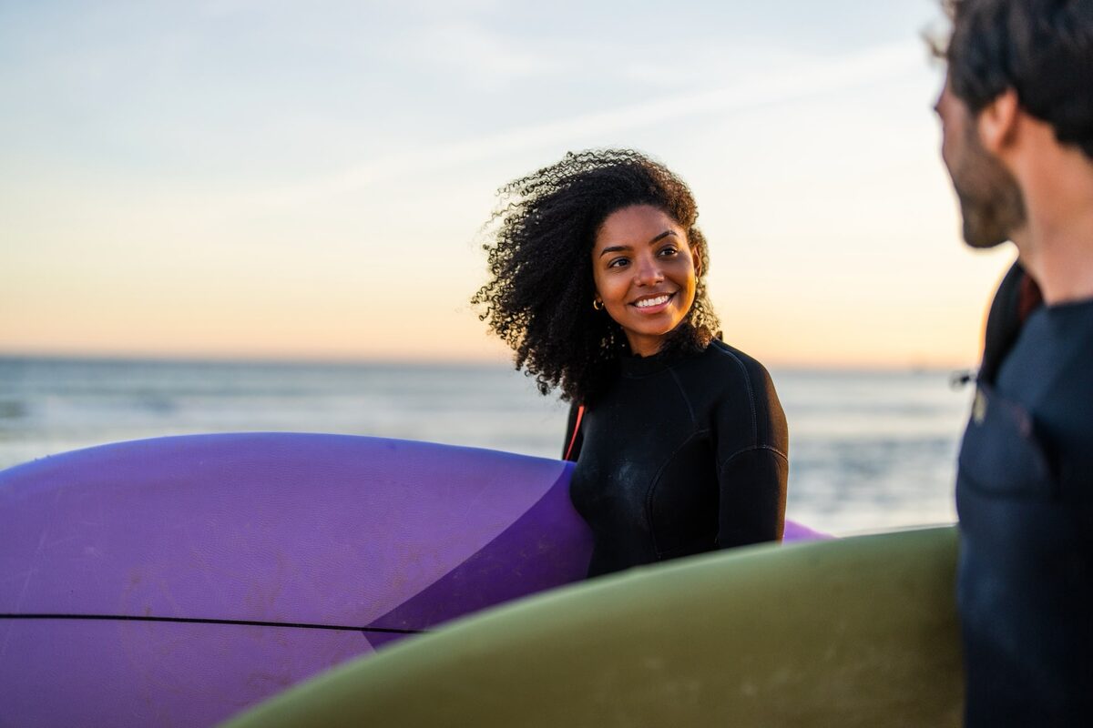 Junge Frau mit Surfbrett am Strand, im Vordergrund ist ein junger Mann von hinten sichtbar.