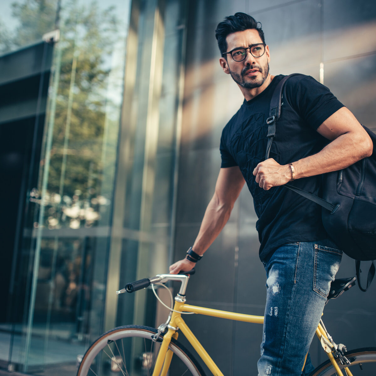 Sportlicher Mann mit Brille und Rucksack vor einem modernen Gebäude. Er schiebt ein Fahrrad.