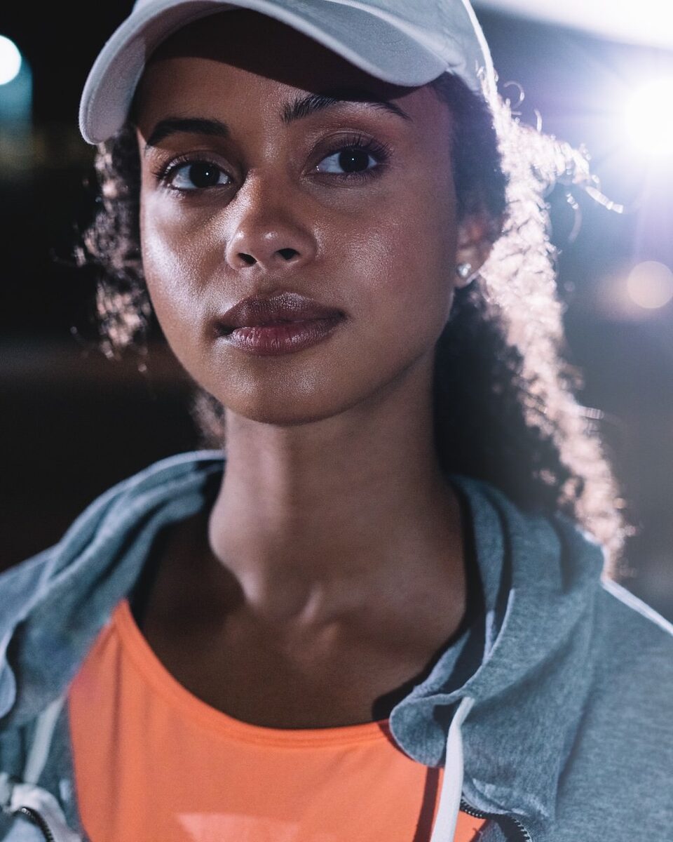 Portrait einer jungen Frau mit Baseballcap bei Flutlicht in einer Sportarena.