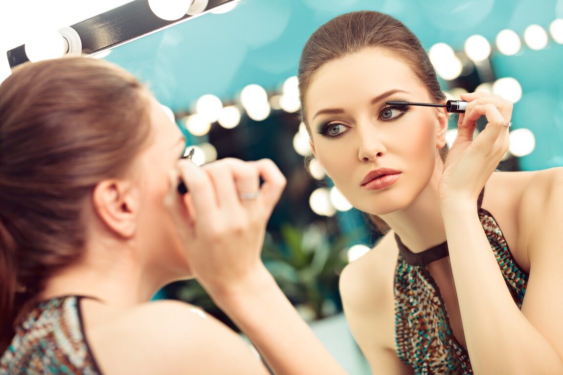 Elegante Frau im Partyoutfit steht vor einem Spiegel und trägt Augen-Make-up auf.