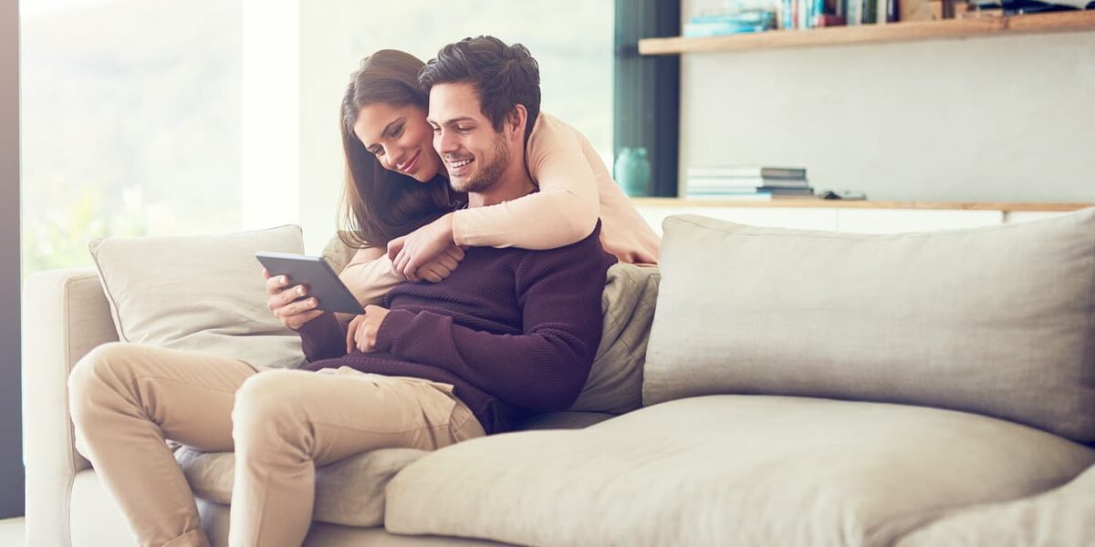 Junges Paar sitzt zuhause auf dem Sofa und schaut in ein iPad.