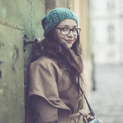 Junge Frau mit Mütze und Brille lehnt an einer Häuserwand und schaut in die Kamera.