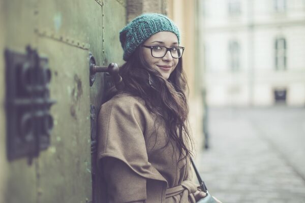 Junge Frau mit Mütze und Brille lehnt an einer Häuserwand und schaut in die Kamera.
