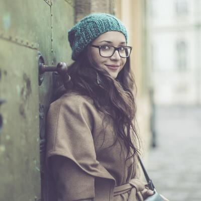 Junge Frau mit Mütze und Brille lehnt an einer Häuserwand und schaut in die Kamera.