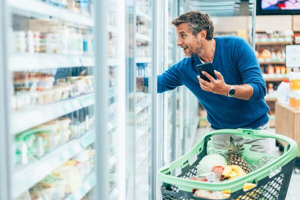 Mann um die 60 kauft ohne Brille im Supermarkt ein. Er hält sein Handy in der Hand.