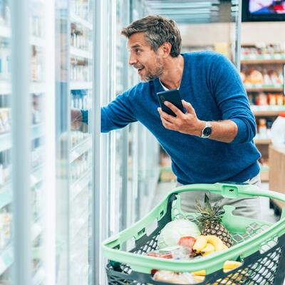 Mann um die 60 kauft ohne Brille im Supermarkt ein. Er hält sein Handy in der Hand.